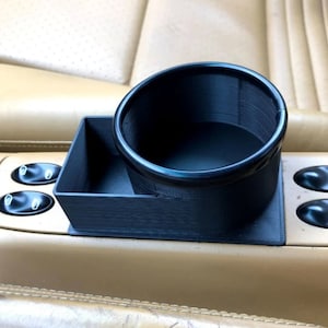 Może przedstawiać: Czarny, wydrukowany w 3D uchwyt na kubek z małym, prostokątnym schowkiem. Uchwyt na kubek jest zamontowany w konsoli środkowej samochodu.