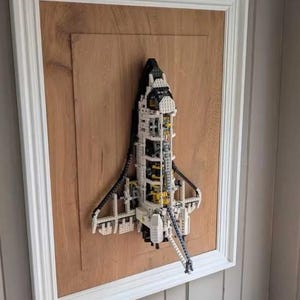 Può includere: Modello di navetta spaziale realizzato con mattoncini bianchi, neri e gialli, incorniciato in bianco. Il modello è montato su un pannello di legno. L'opera d'arte è appesa a una parete grigia, creando un effetto 3D.