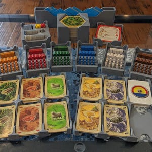 May include: A board game organizer set with various components. The set includes gray plastic trays holding wooden pieces in multiple colors, dice, and cards with illustrated landscapes. The game pieces are arranged on a reflective surface.