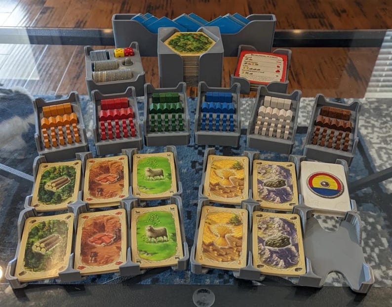 May include: A board game organizer set with various components. The set includes gray plastic trays holding wooden pieces in multiple colors, dice, and cards with illustrated landscapes. The game pieces are arranged on a reflective surface.
