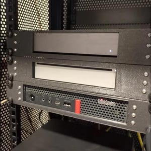 Puede incluir: Un rack de servidor negro con tres estantes metálicos. El estante inferior tiene una etiqueta negra ThinkCentre y varios puertos. Los dos estantes superiores tienen un panel frontal negro con un borde plateado.