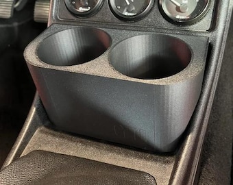 May include: Black 3D-printed car console with two cup holders and three gauges. The gauges display oil pressure, oil temperature, and voltage. The console is designed to fit in a car's center console area.