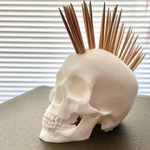 May include: A white skull-shaped container with wooden toothpicks arranged to resemble a mohawk. The container is made of a smooth, matte material and has detailed facial features. The toothpicks are a light brown colour.