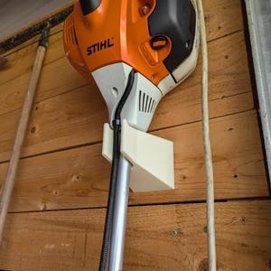 May include: An orange and black STIHL string trimmer is mounted on a wooden wall. The trimmer is held in place by a cream-colored bracket. The wooden wall has a natural wood grain pattern.