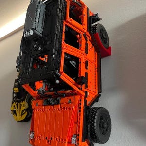 Könnte beinhalten: Ein orange-schwarzes Modell eines Fahrzeugs, das aus ineinandergreifenden Bausteinen besteht und an einer Wand befestigt ist. Das Modell verfügt über große schwarze Reifen und komplizierte mechanische Details. Das Fahrzeug ist leuchtend orange.