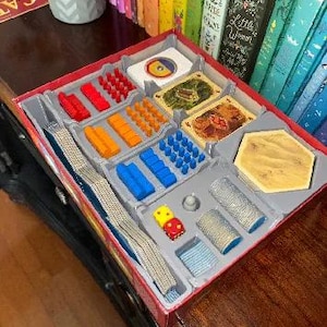 May include: A board game box with various game pieces, including red, orange, and blue game tokens, cards, dice, and hexagonal tiles. The box is red with a gray interior, and the game pieces are neatly organized.