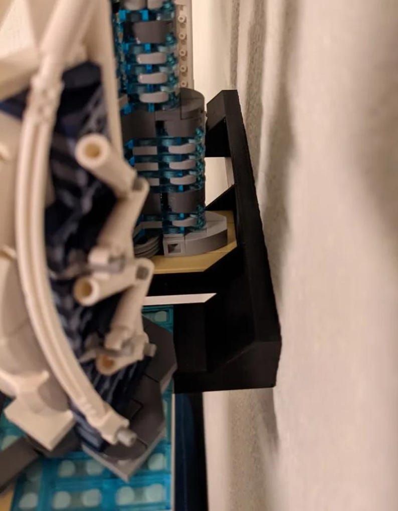 Puede incluir: Un soporte de pared de pl&aacute;stico negro para exhibir un modelo de Lego. El soporte est&aacute; dise&ntilde;ado para sujetar el modelo de forma segura a la pared.
