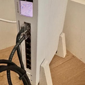 以下が含まれることがあります： パターンを表示する小さな画面を備えた白いネットワークデバイス。黒いケーブルがデバイスに接続されており、2つの白いスタンドで支えられています。デバイスは木製の表面にあります。