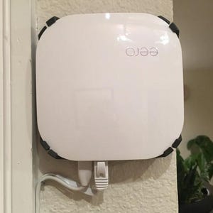 May include: A white square-shaped eero wifi router mounted on a wall with black corner brackets. The router has a white power cord plugged into the bottom.