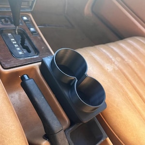 以下が含まれることがあります： 車用の黒いデュアルカップホルダー。センターコンソールに配置されています。カップホルダーはプラスチック製で、パーキングブレーキとギアシフトの隣にあります。インテリアはタン色の革張りのシートと木製のトリムです。