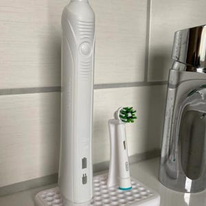Pode incluir: Uma escova de dentes elétrica branca e uma cabeça de escova de reposição em um suporte de carregamento de plástico branco com um design de favo de mel.