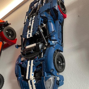 May include: A blue and white model car, mounted on a wall. The car has black wheels and white racing stripes. The model is a detailed replica of a sports car. Another orange model car is partially visible in the background.