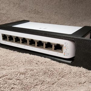 Op de afbeelding: Een witte netwerkswitch met acht poorten, gehuisvest in een zwart rackmount. De switch heeft een witte bovenkant met het "U" logo en een rij indicatielampjes. De rackmount heeft montagegaten.