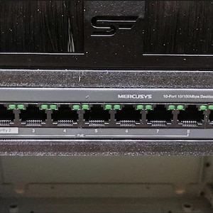 Puede incluir: Un dispositivo de red MERCUSYS de 10 puertos 10/100 Mbps para escritorio con 8 puertos PoE+ de color negro y gris. El dispositivo tiene diez puertos Ethernet con indicadores verdes y está montado en un bastidor negro.