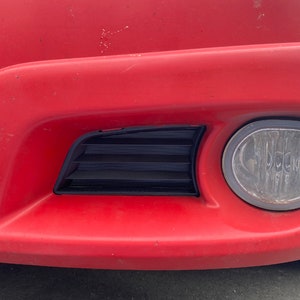 Peut inclure: Un pare-chocs de voiture rouge avec une grille en plastique noire et un feu antibrouillard rond.