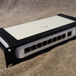 Op de afbeelding: Een wit en zwart netwerkswitch met meerdere Ethernet-poorten. Het apparaat is gemonteerd in een zwart rek en heeft het logo van het merk aan de bovenkant. De switch is ontworpen voor netwerkconnectiviteit en gegevensoverdracht.