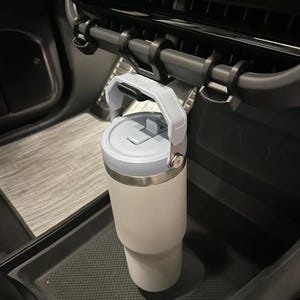 May include: A gray and white insulated travel mug with a black handle is hanging from a black car cup holder.