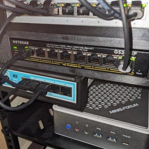 Op de afbeelding: Een netwerkrek met meerdere elektronische apparaten, waaronder een NETGEAR GS316P switch, een kleinere Hex switch en een MINIS FORUM computer. Kabels verbinden de apparaten en het rek is aan een muur bevestigd.