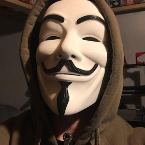 May include: A white Anonymous mask with black eyebrows, mustache, and goatee. The mask is worn with a brown hooded sweatshirt. The mask has a smiling expression.