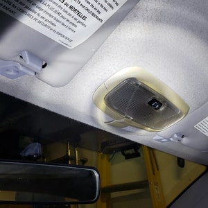 May include: A white overhead light fixture in a car with a grey fabric headliner. The light fixture has a clear plastic cover and a small button on the side. The light is on and illuminating the interior of the car.