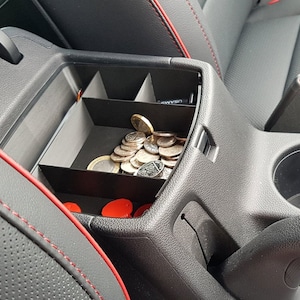May include: A black car centre console with a red trim. The console has a small storage compartment with a black divider and a cup holder. There are several coins in the storage compartment.