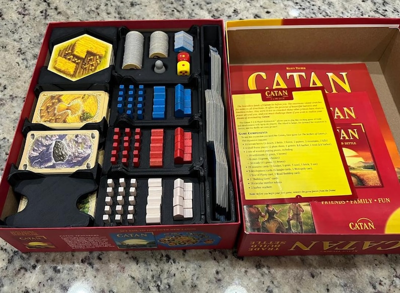 May include: A board game set with various game pieces, including cards, dice, and wooden components, arranged in a red box. The box is open, revealing the game's contents, with the game title "CATAN" visible on the box and components.