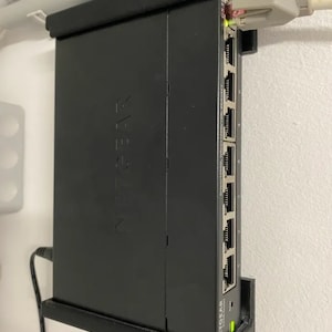 Puede incluir: Un conmutador Netgear negro montado en una pared con un cable blanco conectado a uno de los puertos.