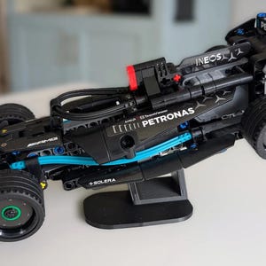 Puede incluir: Un modelo de coche de carreras negro y gris con detalles azules y rojos, expuesto sobre un soporte negro. El coche presenta los logotipos "PETRONAS" e "INEOS", y tiene ruedas negras con centros verdes. El modelo es una réplica detallada.