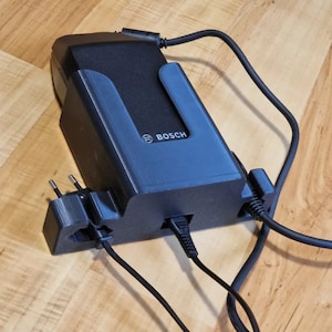May include: A black Bosch power tool battery charger with a wall adapter and a power cord. The charger has a built-in holder for the battery.