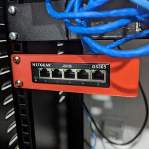 Może przedstawiać: Czerwony przełącznik sieciowy Netgear GS305 zamontowany w szafie serwerowej. Przełącznik posiada pięć portów Ethernet z zielonymi diodami aktywności. Niebieskie kable sieciowe są widoczne nad przełącznikiem.