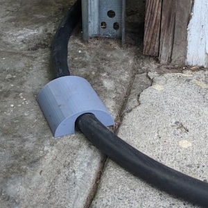 May include: A grey, semi-circular cable protector made of plastic. The protector is placed over a black cable, likely an electrical cord, to prevent damage. The cable protector is on a concrete surface.