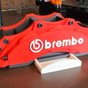 May include: A red Brembo brake caliper with black accents and the Brembo logo in white. A small white tray with the Brembo logo is in front of the caliper. The caliper is likely a car part.