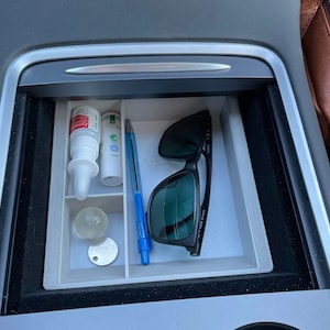 Puede incluir: Una bandeja organizadora de plástico blanco con compartimentos para guardar objetos pequeños. La bandeja contiene un bolígrafo azul, una botella transparente, un pequeño tubo blanco, un objeto redondo transparente, un llavero plateado y unas gafas de sol negras.