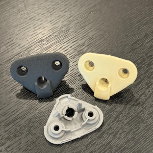 May include: Three plastic car parts in different colours: black, beige, and grey. The parts are triangular in shape with three holes and a central hole.
