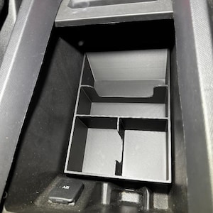 May include: Black plastic car console organizer with multiple compartments. The organizer is designed to fit inside a car's center console, providing storage for various items. The 12V power outlet is visible.