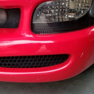 May include: Close-up of a red car's front bumper with a black honeycomb grille and a clear headlight.