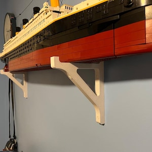 Puede incluir: Un soporte de estante de madera blanco sostiene un gran modelo del RMS Titanic, hecho de ladrillos LEGO. El barco está pintado de rojo, negro y amarillo.