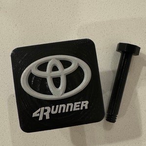 May include: A black square-shaped key chain with a silver Toyota logo and the text "4RUNNER" in silver lettering. A black screw is included.