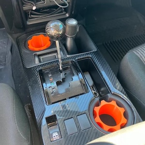 May include: Interior car console with a carbon fiber pattern. Features a gear shift with a black and silver ball, an orange cup holder insert, and an "AUTO" button.