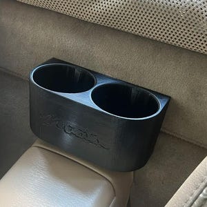 May include: Black plastic cup holder with two slots for beverages. The holder is attached to the back of a car seat.