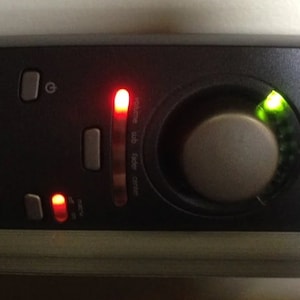 May include: A black audio mixer with a volume knob, a red light, and a green light. The mixer has a microphone input and a headphone output.