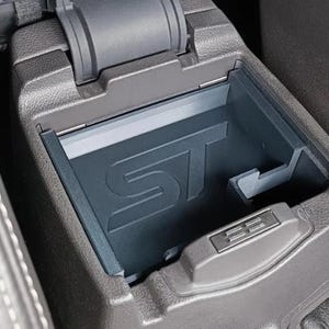 May include: A dark gray car console storage tray with the letters "ST" embossed on the bottom. The tray is open, revealing the interior. A small, rectangular button is visible on the front edge of the tray.