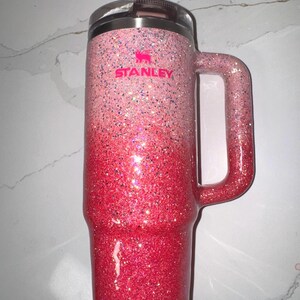 Może przedstawiać: R&oacute;żowy kubek Stanley z brokatem, z gradientem kolor&oacute;w od jasnor&oacute;żowego do ciemnoczerwonego. Posiada uchwyt i logo Stanley w kolorze r&oacute;żowym.