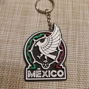 Puede incluir: Un llavero negro y blanco con un anillo de llave plateado. El llavero presenta una imagen estilizada del escudo de armas mexicano, con un esquema de colores verde, blanco y rojo. La palabra "MEXICO" está impresa en letras blancas en el llavero.