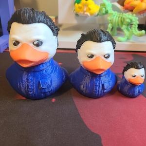 May include: Three rubber duck bath toys in the shape of a person with black hair and a blue jumpsuit. The ducks are different sizes, with the largest duck in the front and the smallest duck in the back.