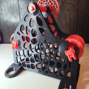 May include: A 3D-printed, black and red mechanical device with a triangular frame and circular cutouts. The device features red gears, rollers, and fasteners. The base has two curved, circular openings. The design is intricate and geometric.