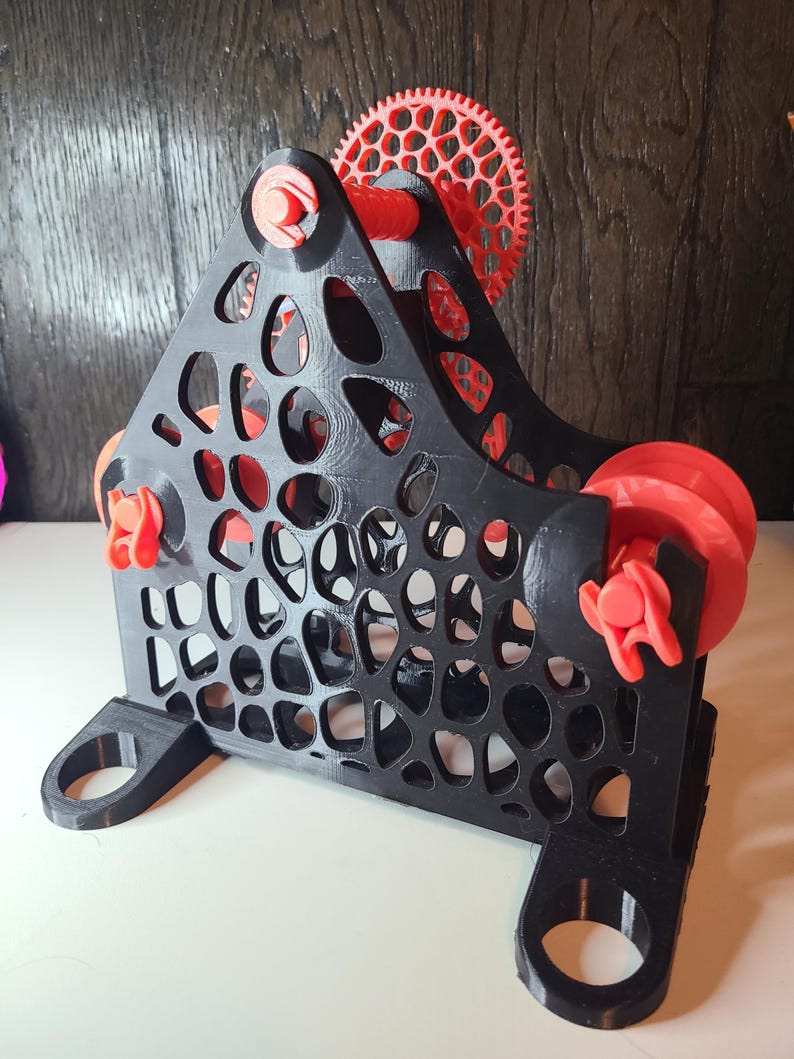 May include: A 3D-printed, black and red mechanical device with a triangular frame and circular cutouts. The device features red gears, rollers, and fasteners. The base has two curved, circular openings. The design is intricate and geometric.