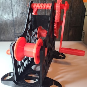 May include: A 3D-printed yarn swift in black and red. The yarn swift has a black frame with a honeycomb pattern, red spools, and a red gear system with a hand crank. The swift is designed to hold yarn for winding.