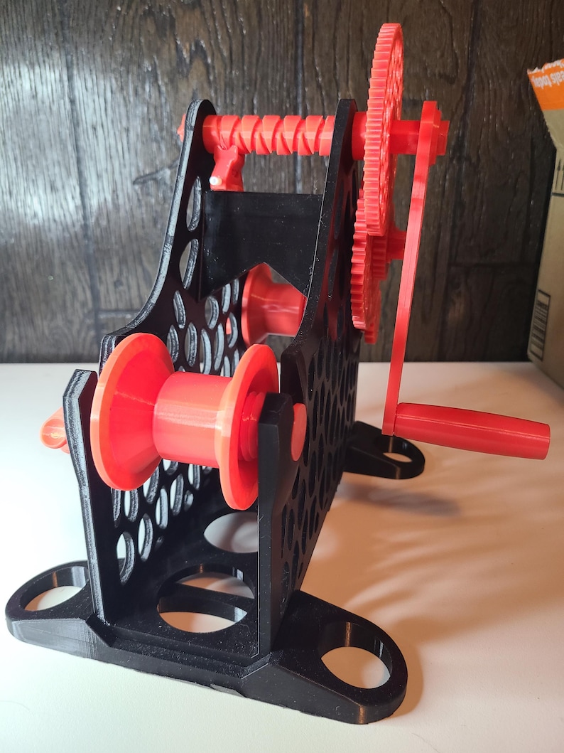 May include: A 3D-printed yarn swift in black and red. The yarn swift has a black frame with a honeycomb pattern, red spools, and a red gear system with a hand crank. The swift is designed to hold yarn for winding.