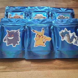 May include: Several blue iridescent pouches with various cartoon character stickers. The stickers include a yellow Pikachu, a purple Gengar, and a blue and white Gyarados. The pouches are sealed and arranged on a wooden surface.